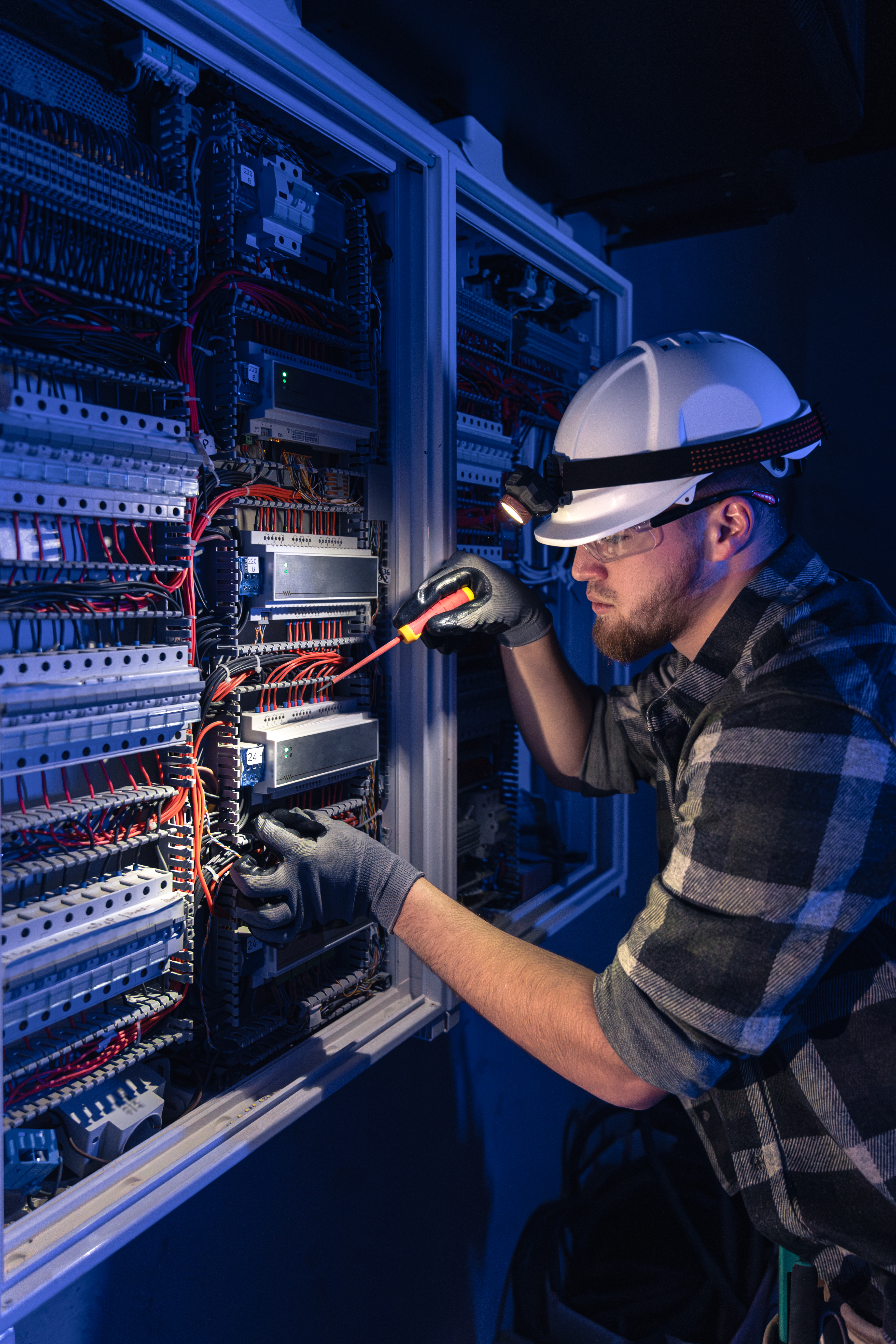 Male electrician working in switchboard with electrical connecting cable. Young adult electrical engineer in special clothes with flashlight on helmet in dark room with emergency lights in background.